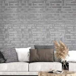 May include: A white couch with gray, brown, and black throw pillows. The couch is in front of a wall with a gray and white patterned wallpaper. A vase with dried pampas grass sits on a wooden coffee table in front of the couch.