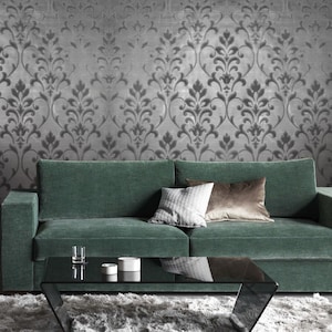 May include: A green velvet sofa with three pillows sits in front of a silver wallpaper with a damask pattern. A glass coffee table sits in front of the sofa.