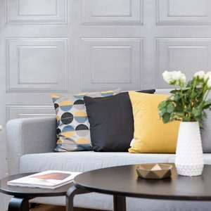 May include: A gray couch with three throw pillows, one with a geometric pattern in blue, yellow, and black, one black, and one yellow. A white vase with white flowers sits on a coffee table in front of the couch.