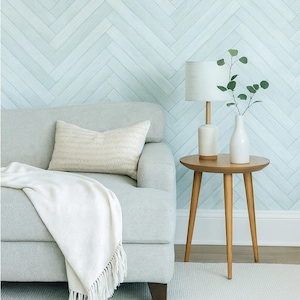 May include: A light gray sofa with a white throw blanket and a textured pillow. A small wooden side table holds a white lamp and a white vase with greenery. The background features a light blue herringbone pattern.