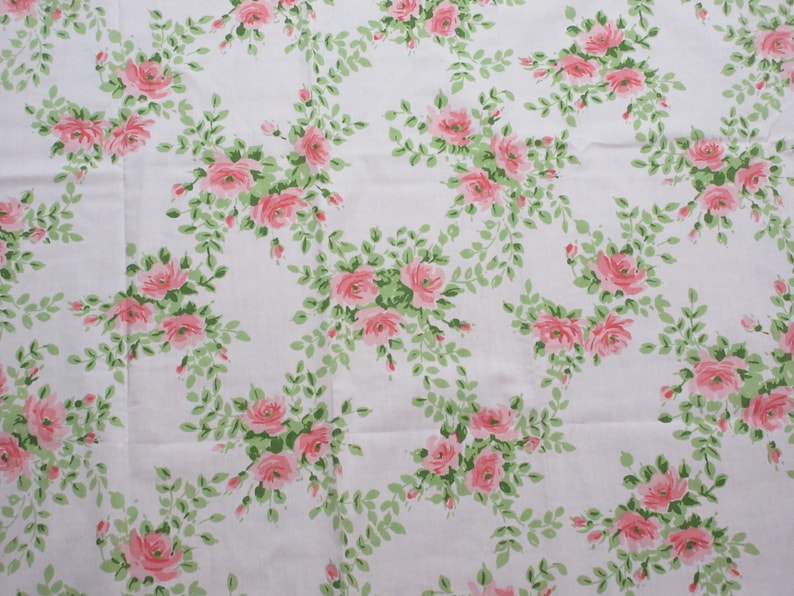May include: White fabric with a repeating pattern of pink roses and green leaves. The roses are clustered in groups, and the leaves form a delicate, winding design across the fabric. The overall effect is a classic, floral print.