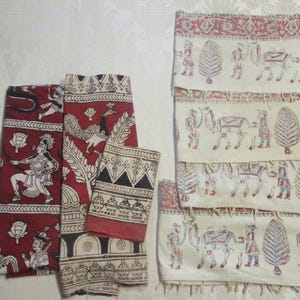 May include: A collection of folded fabric pieces with intricate block-printed designs. The fabrics feature a deep red background with white and black patterns, including figures, animals, and geometric shapes. Some pieces have a cream background with red, blue, and black motifs.