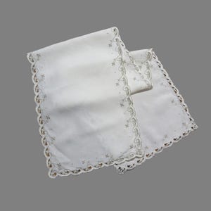 May include: Two white linen table runners with scalloped edges and embroidered floral designs. The runners are folded and layered, showcasing the delicate details of the fabric and embroidery.