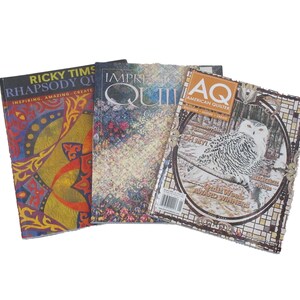 May include: Three quilting magazines are displayed against a white background. The magazines feature colorful covers with titles like "Rhapsody Quilts," "Improvisational Quilts," and "AQ American Quilter." The magazines are for quilting enthusiasts.