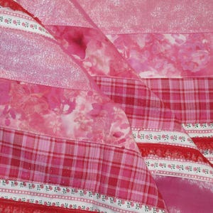 May include: A patchwork quilt featuring various shades of pink fabric. The design includes strips of shimmering pink material, floral patterns, red and white plaid squares, and decorative white ribbon trim with small pink rosebud motifs.