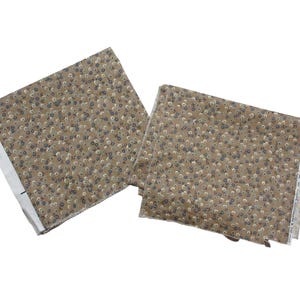 May include: Two pieces of fabric with a floral pattern. The fabric has a tan background with small, scattered flowers in shades of white, blue, and gray. The edges of the fabric are visible, showing a white selvedge.