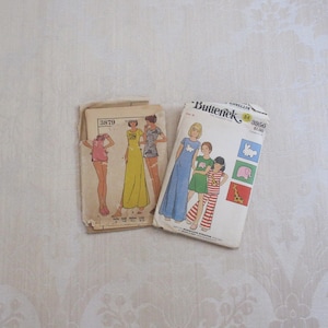 May include: Two vintage sewing pattern packets. One features a woman in a yellow dress, the other shows a woman in a blue dress and children in pajamas. The patterns are on a cream-colored background.