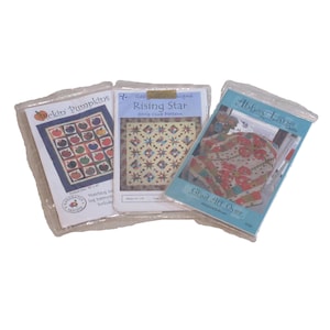 Op de afbeelding: Drie quiltpatroonpakketten. Het patroon "Pickin' Pumpkins" heeft een raster van pompoenen. "Rising Star" heeft een sterrenontwerp. "Glad All Over" toont een bloemenpatroon. Allemaal in doorzichtige plastic verpakkingen.