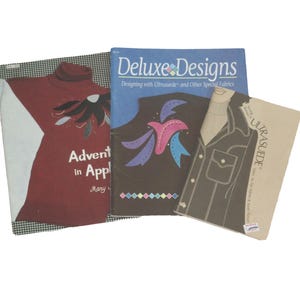 May include: Three vintage sewing pattern books. One book is titled "Advent in Applique" with a red turtleneck design. Another is "Deluxe Designs" with a bird design. The third book is titled "Ultrasuede" with a jacket design.