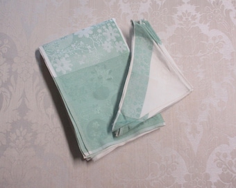 May include: Two folded, light green and white floral patterned cloth napkins. The napkins have a delicate floral design and are folded neatly. The napkins are on a light-colored surface.