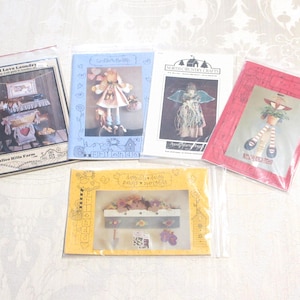 Op de afbeelding: Een verzameling knutselpatronen in plastic hoezen. De patronen tonen verschillende volkskunstontwerpen, waaronder poppen, engelen en wasgoed-thema decoraties. De patronen zijn in verschillende kleuren, waaronder blauw, rood en geel.