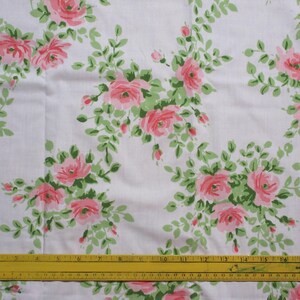 May include: White fabric with a repeating pattern of pink roses and green leaves. The roses are in full bloom and surrounded by smaller buds and foliage. A yellow ruler is at the bottom of the image.