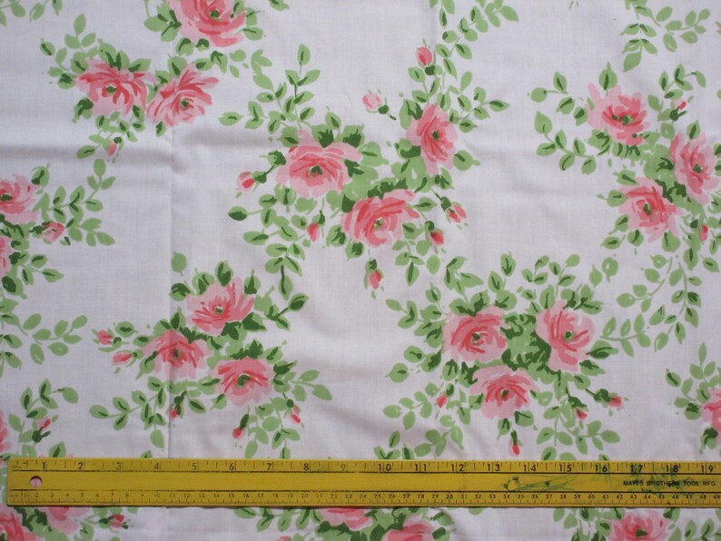 May include: White fabric with a repeating pattern of pink roses and green leaves. The roses are in full bloom and surrounded by smaller buds and foliage. A yellow ruler is at the bottom of the image.