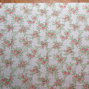 May include: White fabric with a repeating pattern of pink roses and green leaves. The floral design is arranged in a flowing, vine-like pattern across the fabric's surface. The fabric appears to be cotton or a similar material, suitable for various sewing projects.