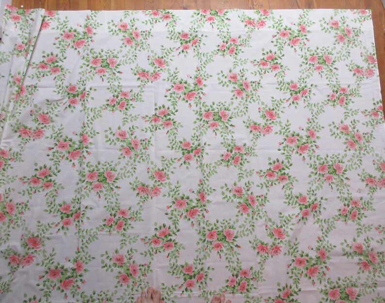 May include: White fabric with a repeating pattern of pink roses and green leaves. The floral design is arranged in a flowing, vine-like pattern across the fabric's surface. The fabric appears to be cotton or a similar material, suitable for various sewing projects.