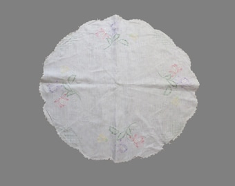 May include: A white, circular doily with a scalloped edge. The doily features embroidered floral designs in pink, purple, and green. The fabric appears slightly wrinkled, suggesting it is made of a soft material.