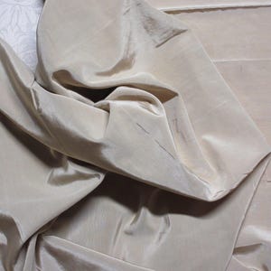 May include: Close-up of a beige fabric with a silky, flowing texture. The fabric has a subtle sheen, with folds and creases creating shadows and highlights. The material appears to be lightweight and drapes softly.