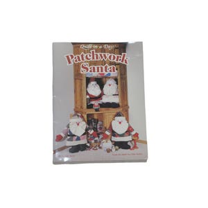 May include: A softcover instructional book titled Quilt in a Day Patchwork Santa. The cover features a photograph of a wooden cabinet displaying four handmade fabric Santa Claus figures with patchwork clothing, white beards, and red hats.