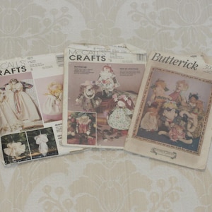 May include: Three vintage sewing pattern packets from McCall's Crafts and Butterick. The packets feature illustrations of dolls and doll clothing. The patterns are on a cream-colored background with a floral design.