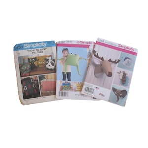 May include: Three Simplicity sewing pattern packets. One packet shows how to sew pillows, another shows a frog pillow, and the third shows an animal head wall decor. The packets are white, blue, and pink.