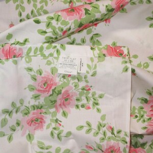 May include: White twin flat sheet with a floral pattern of pink roses and green leaves. The sheet has a tag that reads "Twin Flat 66 x 104". The sheet is made of a soft, likely cotton material.