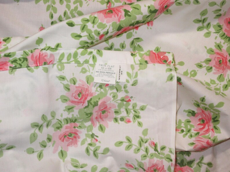 May include: White twin flat sheet with a floral pattern of pink roses and green leaves. The sheet has a tag that reads "Twin Flat 66 x 104". The sheet is made of a soft, likely cotton material.