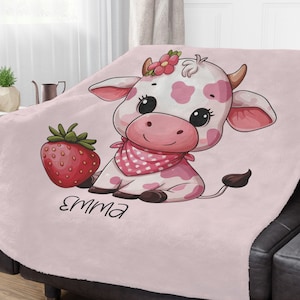May include: Pink fleece blanket with a cartoon cow wearing a pink and white polka dot bandana and holding a red strawberry. The cow has pink spots and a pink flower on its head. The blanket has the name "Emma" written on it.