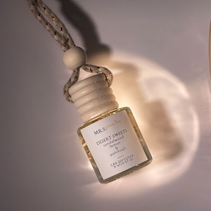 May include: A white car diffuser with a gold string hanging from the top. The bottle is labeled "MRS. CANDLIER DESERT SWEETS sandalwood lemon & patchouli CAR DIFFUSER 8 ml / 0.27 oz"