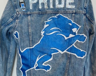 Puede incluir: Una chaqueta vaquera azul con un gráfico de león azul y la palabra "PRIDE" en purpurina plateada en la parte posterior.