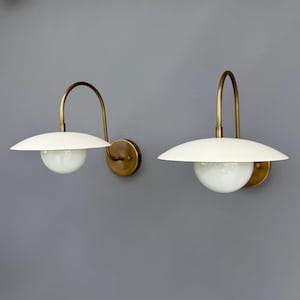 Op de afbeelding: Twee wandkandelaars met een mid-century modern design. Elke kandelaar heeft een witte, schotelvormige kap, een messingkleurige gebogen arm en een ronde, matglazen bol. De wandkandelaars zijn gemonteerd op een messingkleurige cirkelvormige basis.