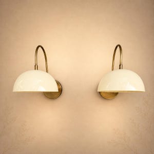 May include: A pair of wall-mounted lamps featuring cream-coloured dome shades and curved brass-toned arms. Each lamp is attached to a circular wall plate, with the dome shade directing light downwards in a classic, functional lighting design.