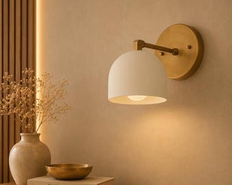 May include: A wall-mounted light fixture featuring a matte white dome-shaped shade attached to a circular brass backplate. The lamp emits a warm glow, highlighting the smooth finish of the metal and the clean lines of the dome design.