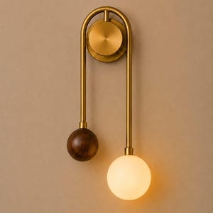 May include: A wall-mounted light fixture featuring a brushed brass frame with a circular backplate. The design includes two vertical rods, one ending in a dark brown wooden sphere and the other in a glowing, spherical white frosted glass shade.