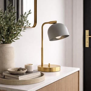 Puede incluir: Una lámpara de mesa con una pantalla gris mate en forma de cúpula y un soporte de metal en tono latón con base circular. La lámpara tiene un brazo vertical sencillo y un pequeño interruptor situado en la parte superior de la base redonda.