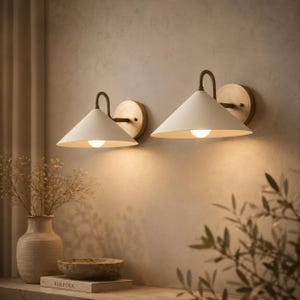 May include: Two wall-mounted lamps featuring matte off-white conical shades and curved brass-toned metal arms. Each lamp is attached to a circular brass-toned wall plate and emits a warm, downward-facing light against the wall surface.