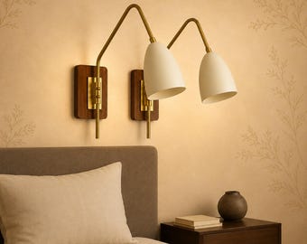 May include: Two wall-mounted lamps featuring matte off-white conical shades, brass-toned metal arms, and rectangular dark wood backplates. The fixtures are positioned side-by-side, each with an adjustable arm extending from the wooden base.