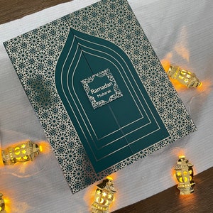 May include: A green and gold Ramadan Mubarak gift box with a decorative geometric pattern. The box has a door-like design with the words "Ramadan Mubarak" on a small label. Gold lantern string lights are arranged around the box.