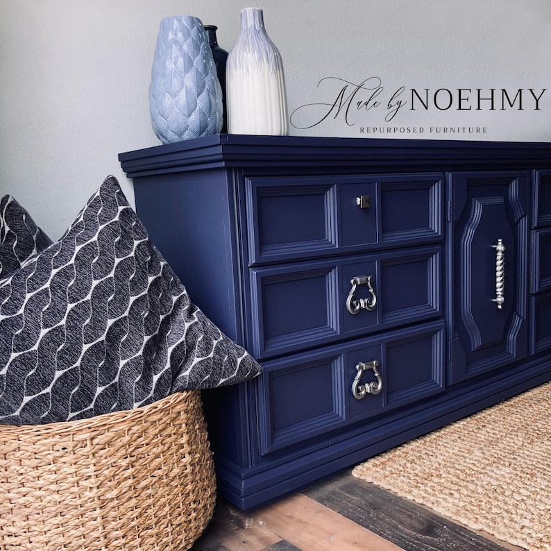 May include: A navy blue dresser with silver hardware, three drawers, and decorative molding. Two decorative vases and a patterned pillow are on top. A woven basket and a jute rug are in the foreground. The text "Made by NOEHMY Repurposed Furniture" is visible.