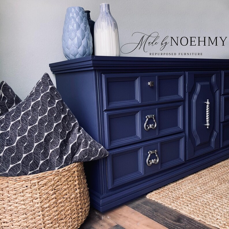 May include: A navy blue dresser with silver hardware, three drawers, and decorative molding. Two decorative vases and a patterned pillow are on top. A woven basket and a jute rug are in the foreground. The text "Made by NOEHMY Repurposed Furniture" is visible.