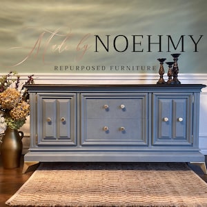 May include: A blue repurposed furniture piece with gold accents. The cabinet has two doors and two drawers with gold and white knobs. The top is dark brown. The words "Made by NOEHMY REPURPOSED FURNITURE" are above the cabinet.