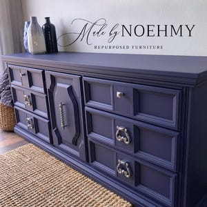 May include: A navy blue repurposed dresser with silver hardware. The dresser has multiple drawers and a central cabinet door with a decorative handle. The text "Made by NOEHMY REPURPOSED FURNITURE" is above the dresser.