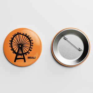 Könnte beinhalten: Orangener Knopf mit einer schwarzen Illustration eines Riesenrads und dem Wort "Miali". Die Rückseite des Knopfes ist silberfarben mit einer Stecknadel.