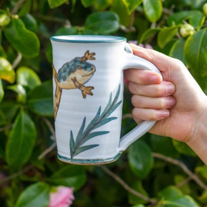 May include: White ceramic mug with a green rim and handle. The mug features a hand-painted design of a green and yellow frog with a leafy branch.