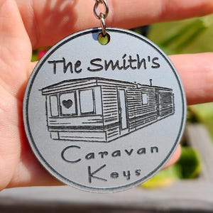 Könnte beinhalten: Ein runder, silberner Schlüsselanhänger mit Kette und Ring. Der Schlüsselanhänger trägt den Text "The Smith's Caravan Keys" und eine Zeichnung eines Wohnwagens. Der Schlüsselanhänger wird in einer Hand gehalten.