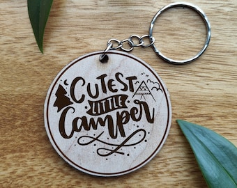 Puede incluir: Un llavero de madera con fondo marrón y texto negro que dice "Cutest Little Camper". El llavero tiene un anillo de metal plateado.