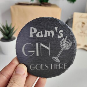 May include: Black slate coaster with white text that reads "Pam's Gin Goes Here". A small etched image of a gin glass with a sprig of rosemary is to the right of the text.