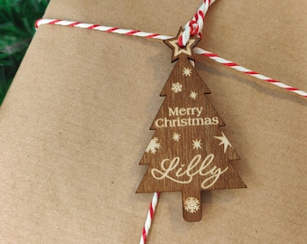 Könnte beinhalten: Eine braune Papiertüte, die mit rotem und weißem Garn gebunden ist. Ein hölzerner Weihnachtsbaum-Anhänger mit dem Text "Merry Christmas Lilly" ist an dem Garn befestigt.