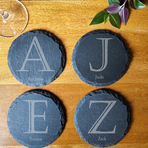 Op de afbeelding: Vier zwarte leisteen onderzetters met een witte letter en een gegraveerde naam op elk. De onderzetters zijn in een vierkant patroon op een houten oppervlak gerangschikt. De letters zijn A, J, E en Z. De namen zijn Andrew, Jude, Emma en Zack.