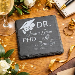 Peut inclure: Un dessous de verre carré en ardoise gris foncé avec du texte et des graphiques blancs. Le texte indique "IT'S DR. Jessica Green PHD Actually! EST. 2025" avec un chapeau de graduation et un diplôme. Le dessous de verre est sur une surface en bois.
