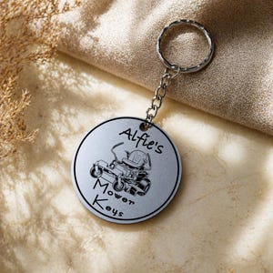 May include: A silver-colored keychain with a round pendant. The pendant features the text "Alfie's Mower Keys" and an illustration of a riding lawn mower. The keychain has a silver ring and chain.